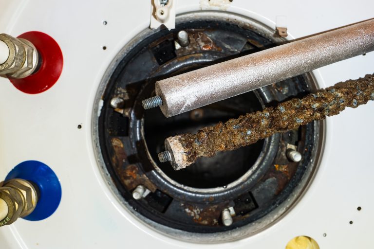 Close-up of two magnesium anodes in front of a water heater tank opening — one new, one corroded — clearly showing wear and protection loss over time.